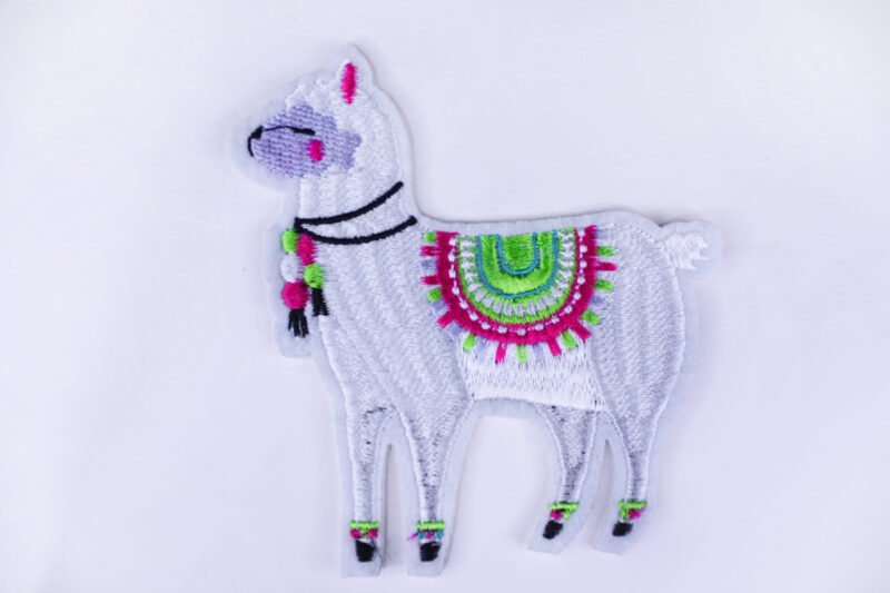 An embroidered patch of a white llama wearing a colourful saddle with tassels and other tasseled accessories.