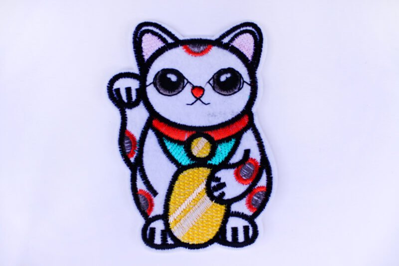 An embroidered patch of a Maneki Neko cat holding a gold disk and waving.