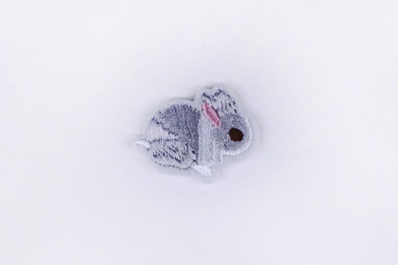 An embroidered patch of a miniature rabbit in the colours grey white and pink.