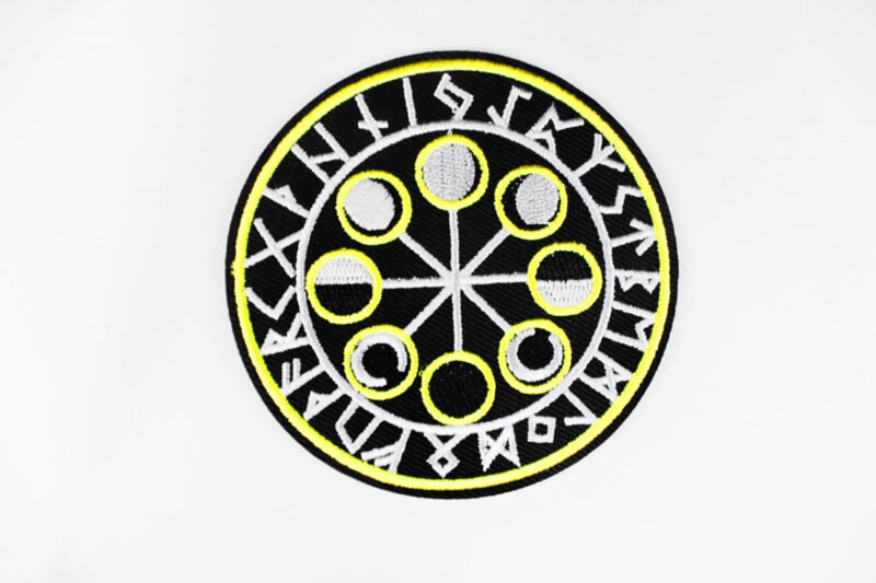 A circular embroidered patch of a the eight moon cycles encircled by Norse runes in black white and yellow.
