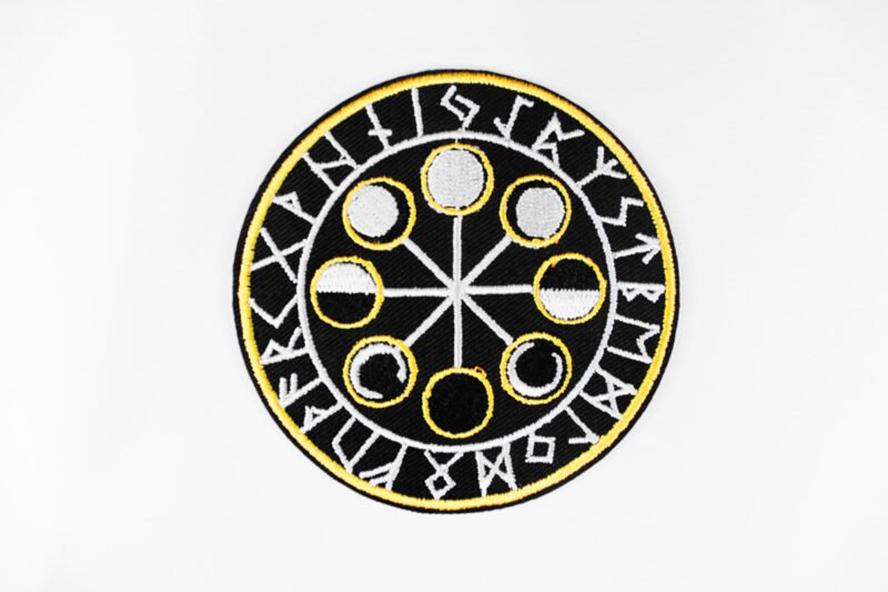A circular embroidered patch of a the eight moon cycles encircled by Norse runes in black white and ochre.