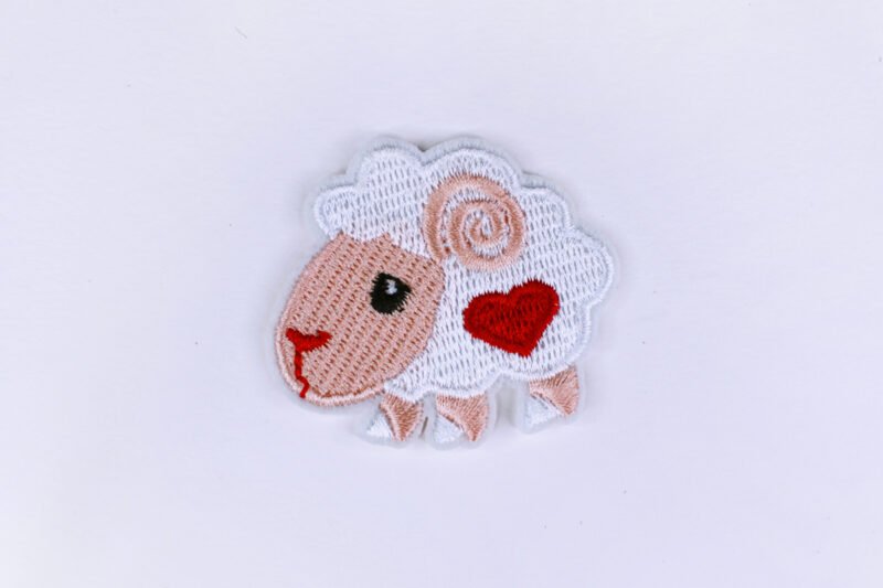 An embroidered patch of a white and peach coloured sheep bearing a red heart on its side.