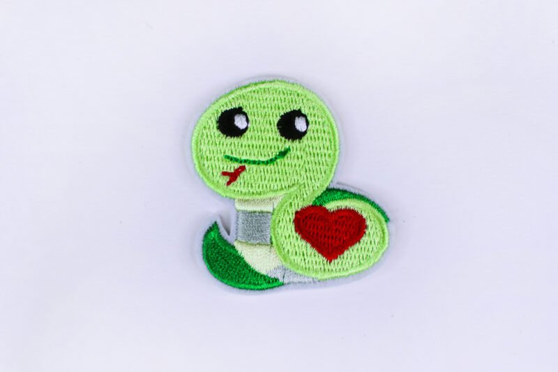 An embroidered patch of a green coloured smiling snake bearing a red coloured heart on its side.