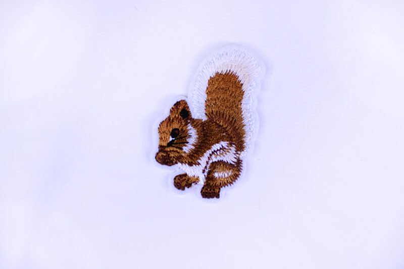 An embroidered patch of a seated brown and white squirrel sniffing its hands.